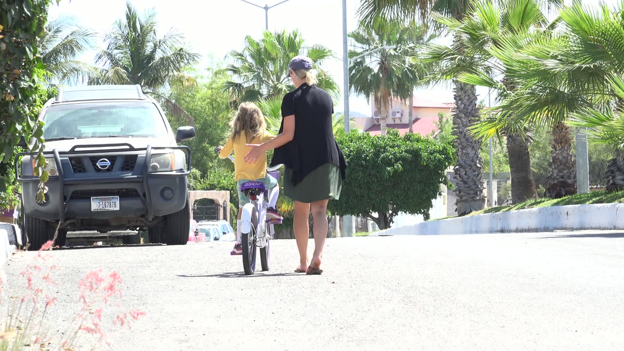 In Los Cabos, Mexico, the child moves slightly ahead on the bicycle while the adult follows close behind, with palms and a parked vehicle lining the street.