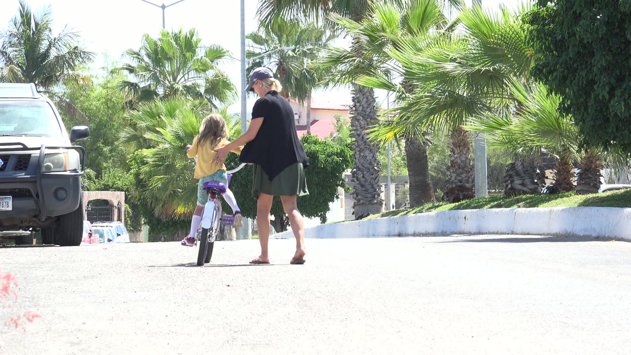 In Los Cabos, Mexico, a woman walks behind and beside a young girl starting to ride a small purple bicycle on a bright paved street with palms and a black SUV nearby.