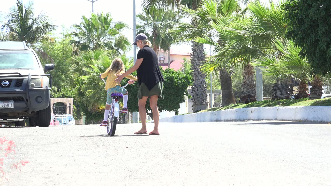 In Los Cabos, Mexico, the young girl pedals forward while the woman keeps a hand near her side, with palm fronds, shrubs, a white wall, and a parked SUV in view.