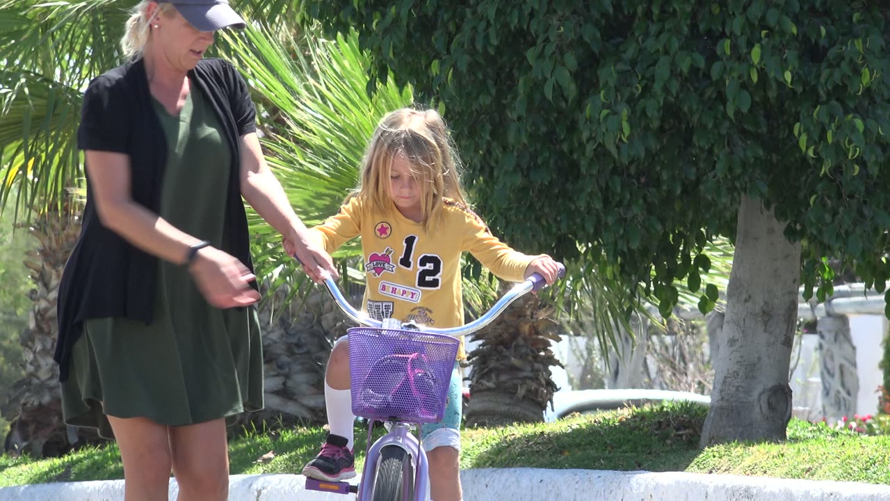 In Los Cabos, Mexico, an adult woman reaches toward a young girl riding a purple bicycle near a low white wall and green landscaping.