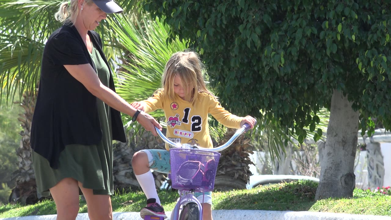 In Los Cabos, Mexico, the girl looks down at the handlebars while the woman stands close with one hand near the bicycle for support.