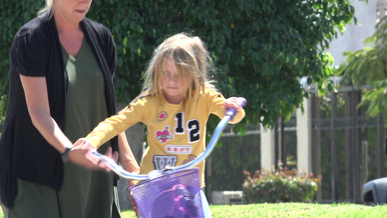 In Los Cabos, Mexico, the woman reaches toward the girl's purple bicycle handlebars while the child looks forward on the grass.