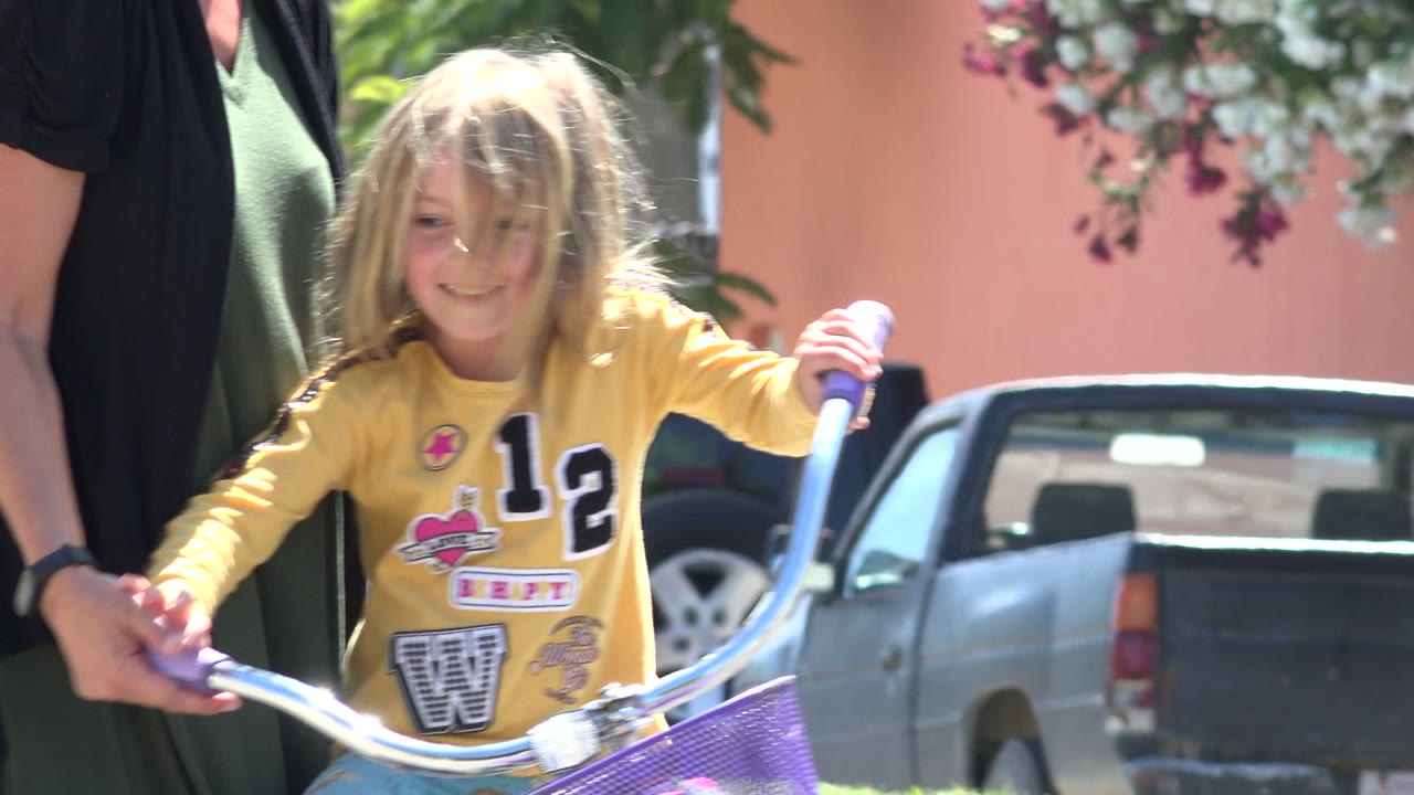 Young blonde child on a bicycle beside an adult on a sunny street.