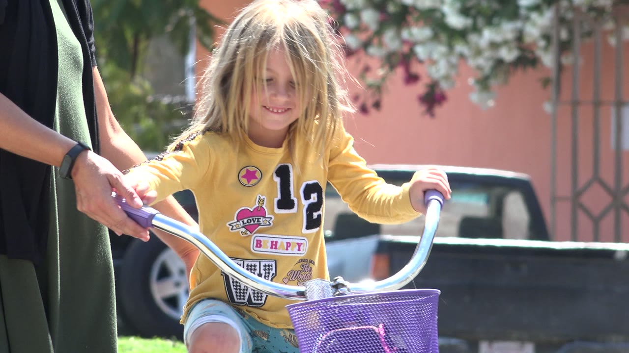 In Los Cabos, Mexico, the child is centered over the bicycle with hands on purple grips while an adult's hand steadies the handlebar in front of a parked pickup and flowering plants.