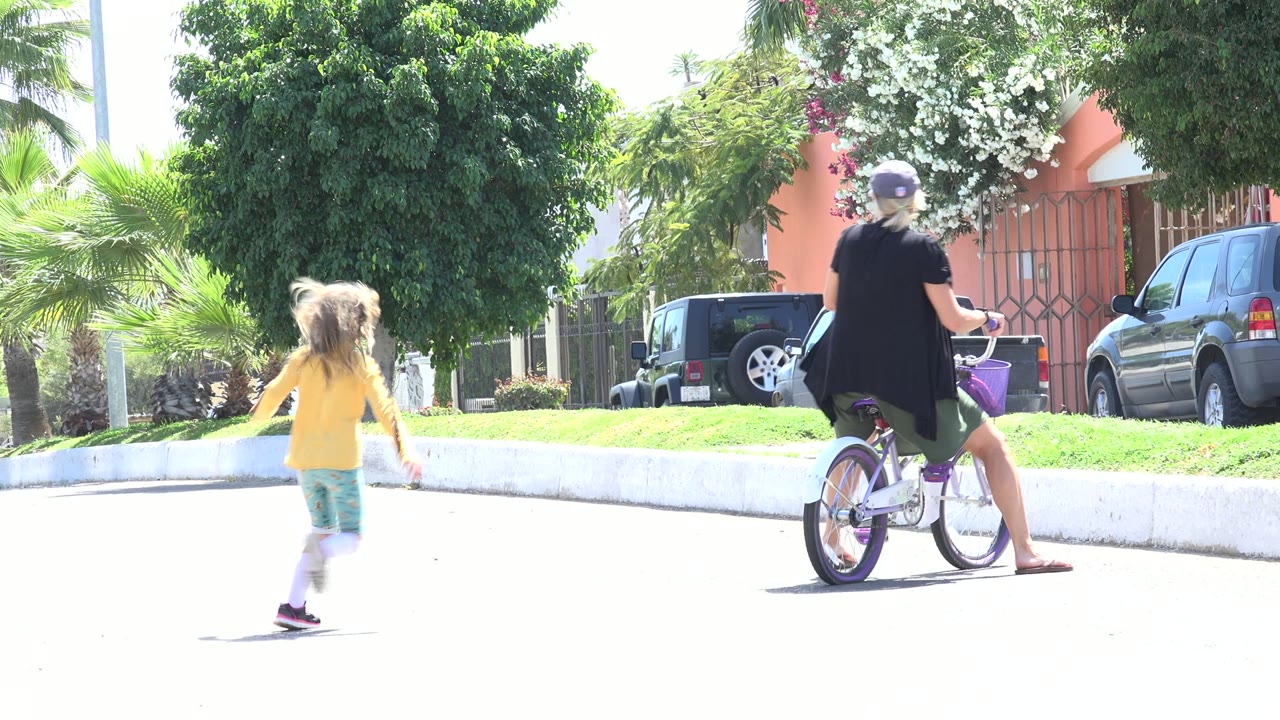 At 0:01 in Los Cabos, Mexico, the girl moves closer to the adult on the purple bicycle, with a white curb, green grass, parked cars, and flowering trees behind them.