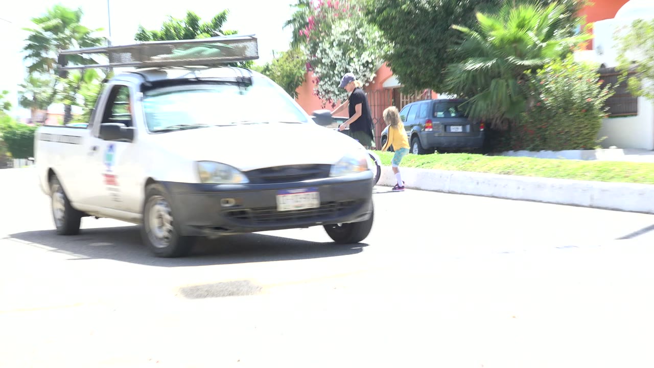 A white utility pickup passes near the foreground on a bright residential street in Los Cabos, Mexico, with an adult and child partially visible near a small bicycle behind it.