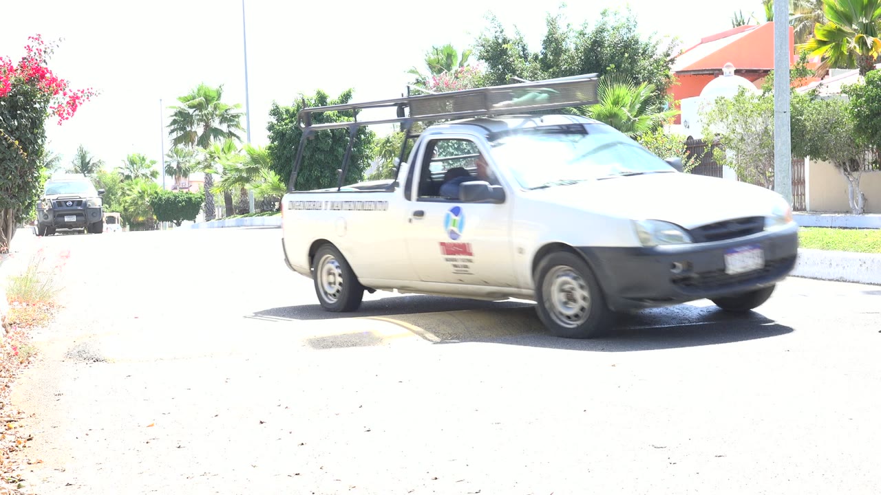 The white pickup moves across the foreground in Los Cabos, Mexico, while the adult and child with a small bicycle are mostly hidden near the right side of the street.