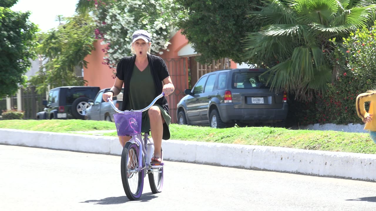 One second in, the woman pedals the purple bicycle on the bright Los Cabos street as the child in yellow moves partly out of frame near parked cars, palms, and a peach-colored building.