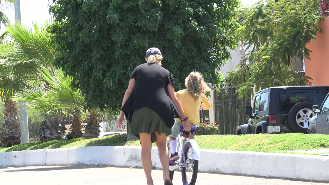 The woman and child move farther along the Los Cabos street, with the adult's hand near the bicycle seat and parked vehicles visible behind them.