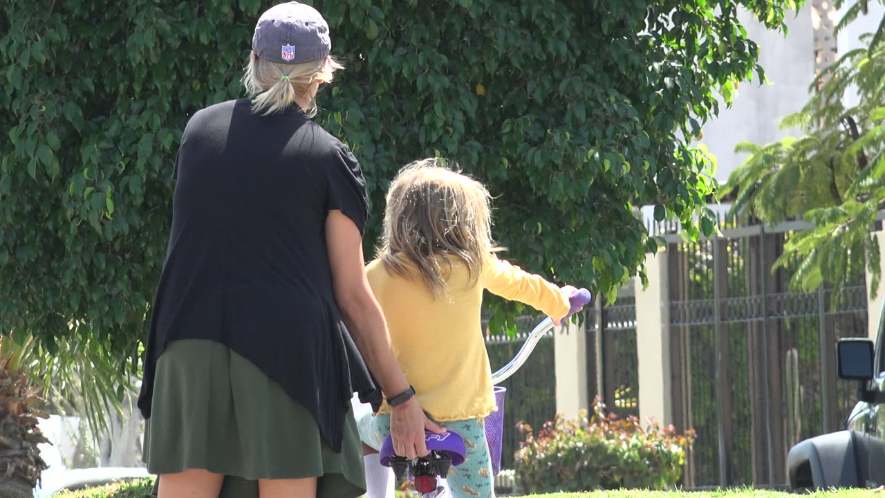 Woman walking behind a young girl on a small bicycle under roadside trees.