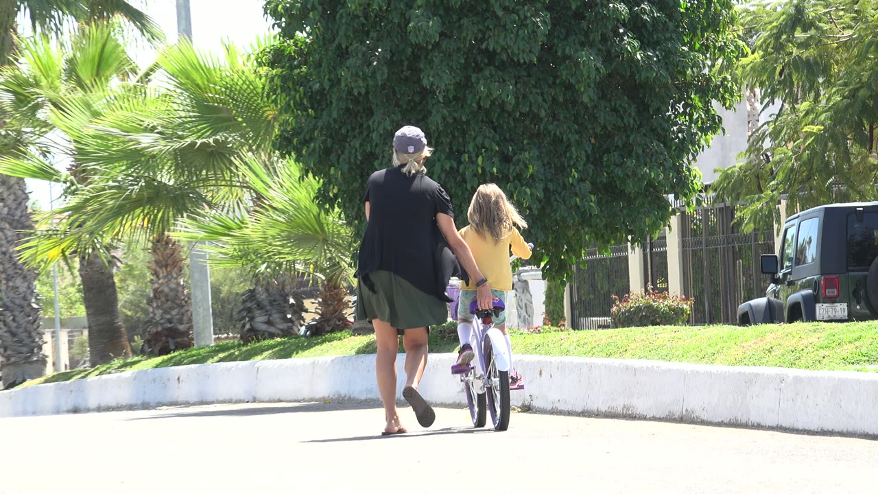 The woman remains close to the child on the small bicycle as they pass a white curb and tropical planting in Los Cabos, Mexico.