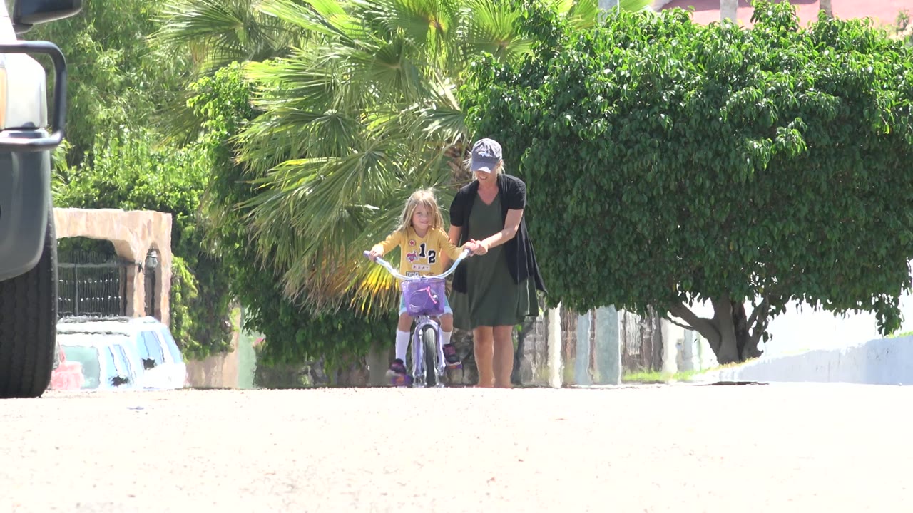 Mother walking beside a young girl on a bicycle in a sunny residential street.