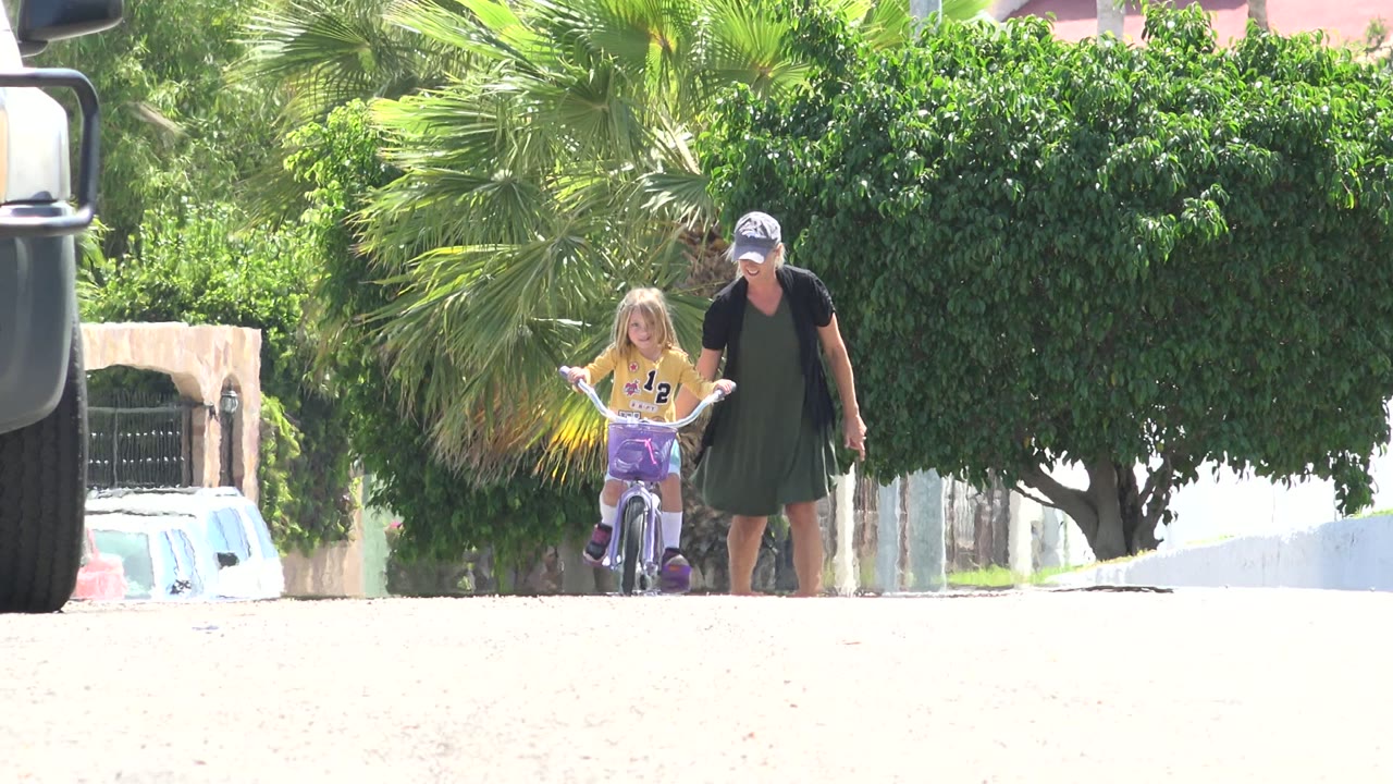 The girl moves forward on her bicycle in Los Cabos while the woman stays at her side, framed by a large green tree, palm leaves, white walls, and parked cars.