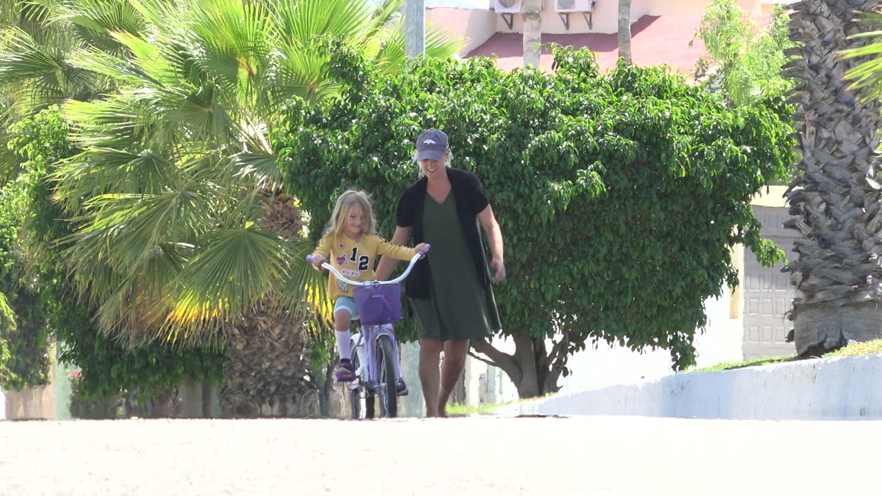 Young girl rides a purple bicycle while a woman walks beside her on a bright street.