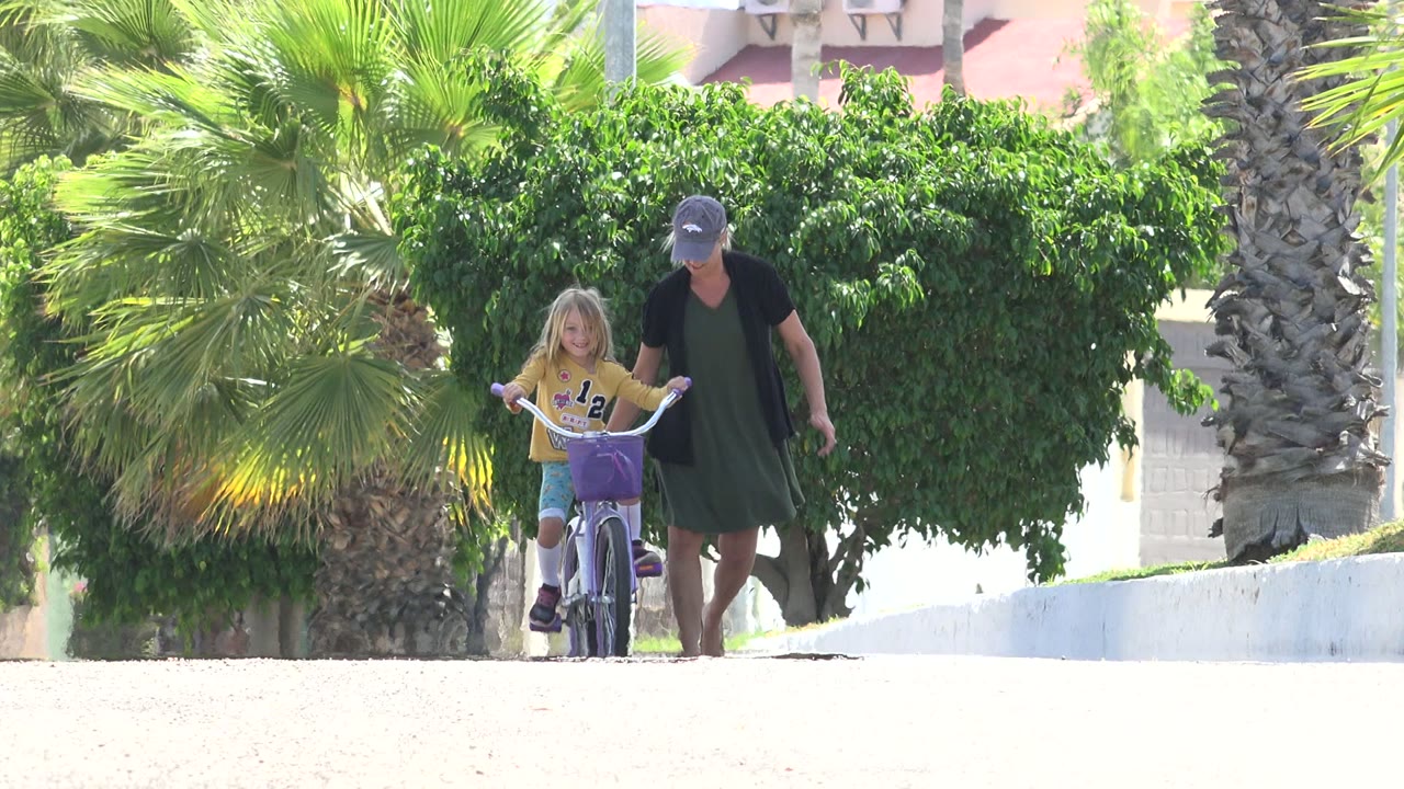 One second in, the girl remains centered on the purple bicycle while the woman walks just to her side, framed by palm leaves, a leafy tree, and bright pavement in Los Cabos, Mexico.