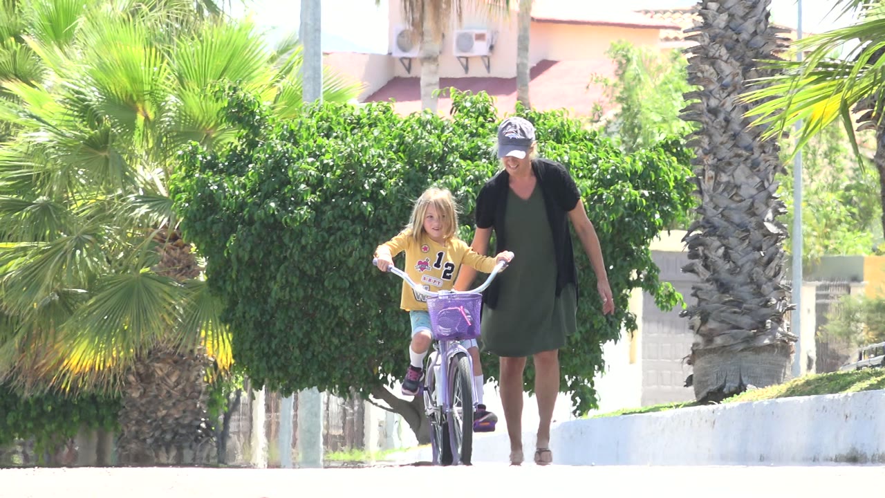 In Los Cabos, Mexico, a woman in a cap walks beside a child riding a purple bicycle along a bright path bordered by palms and trimmed greenery.