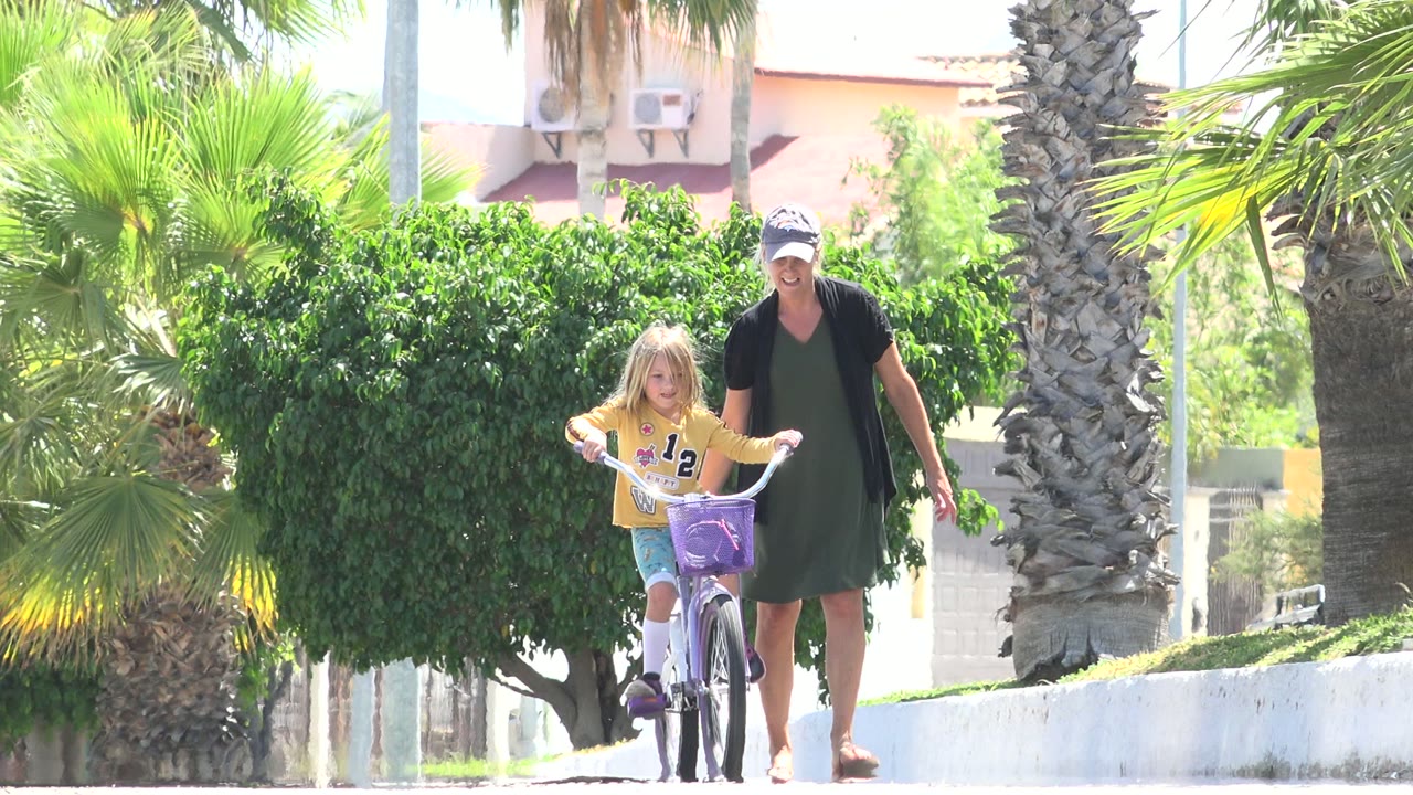 In Los Cabos, Mexico, the child continues forward on the purple bicycle while the woman stays close behind near a palm-lined walkway.