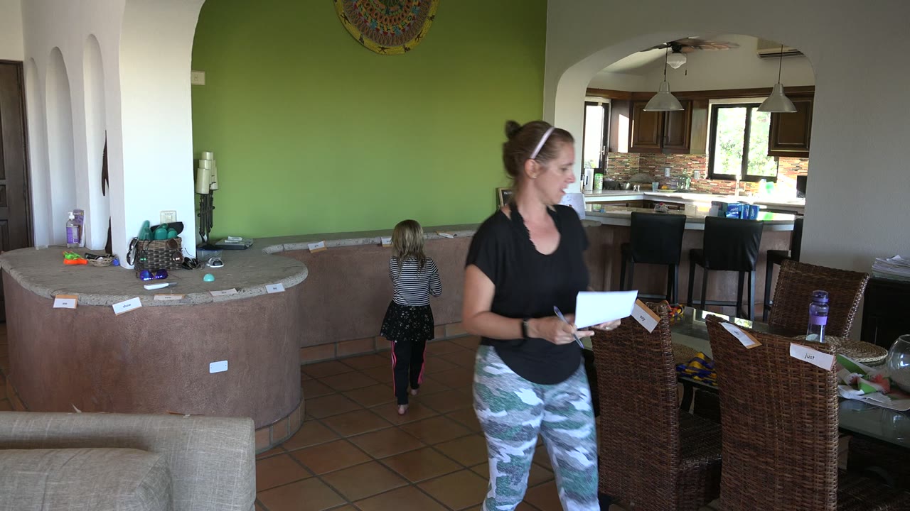 In the Los Cabos dining room, the woman holds papers in the foreground while the young girl faces away near the green wall and the kitchen remains visible in back.