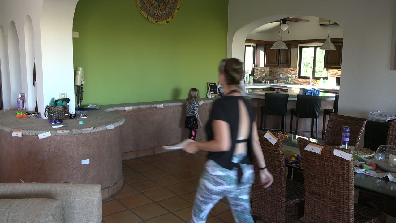 Woman walks through a home interior while a child stands near the kitchen opening.