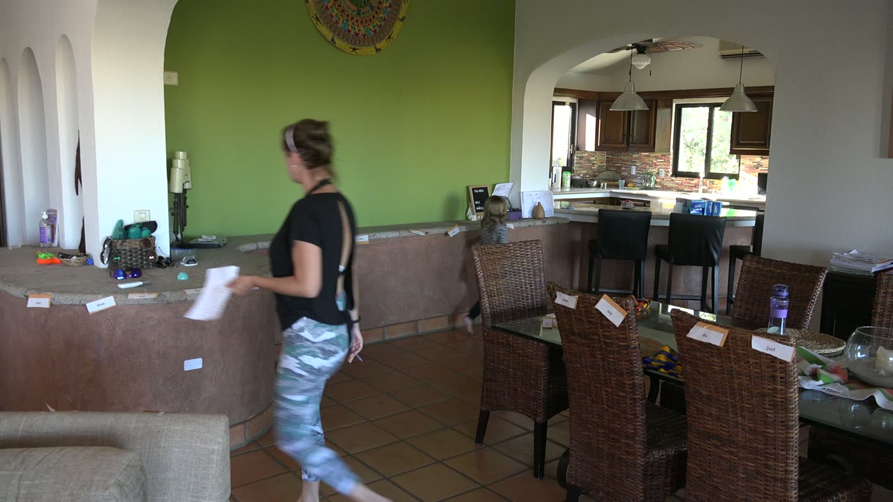 At 0:01 in the Los Cabos interior, the woman stands near the curved counter facing the green wall as the child moves at the kitchen threshold.