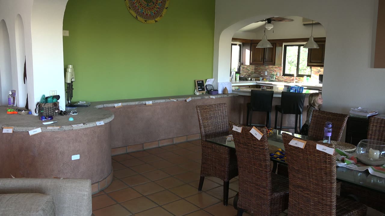 Wide Los Cabos home interior at the start of the clip, with wicker dining chairs in front, a curved counter, a green wall, and a child visible near the kitchen opening.
