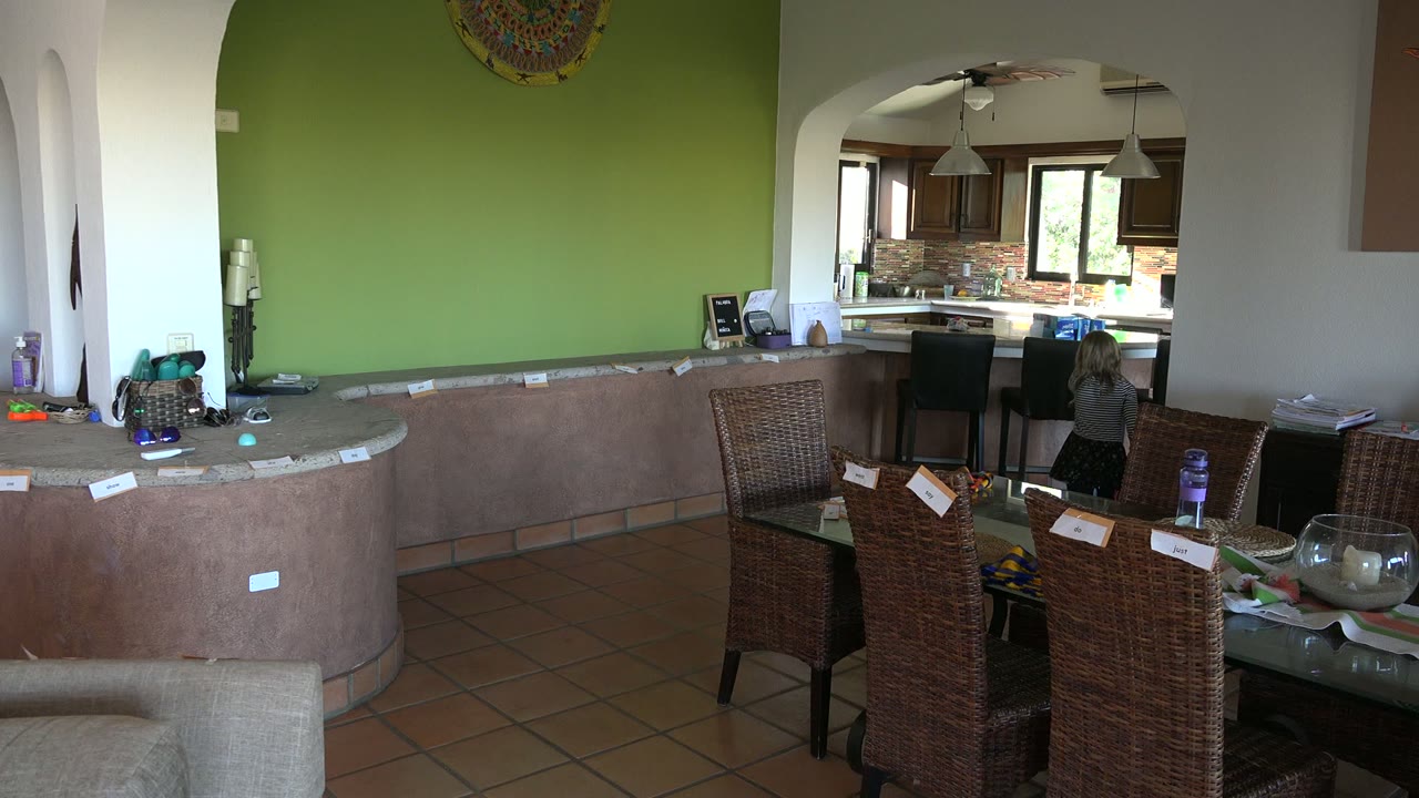 Los Cabos home interior one second in, with the child closer to the kitchen arch, wicker chairs and a glass table in the foreground, and household items along the counter.