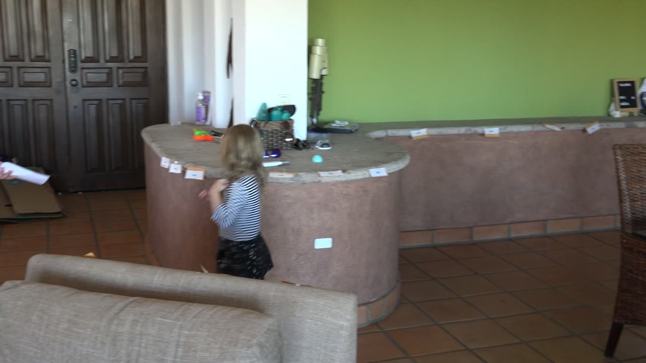 In Los Cabos, Mexico, a blonde child in a striped shirt stands near a curved counter with paper clues while an adult woman is partly visible at the edge of the indoor room.