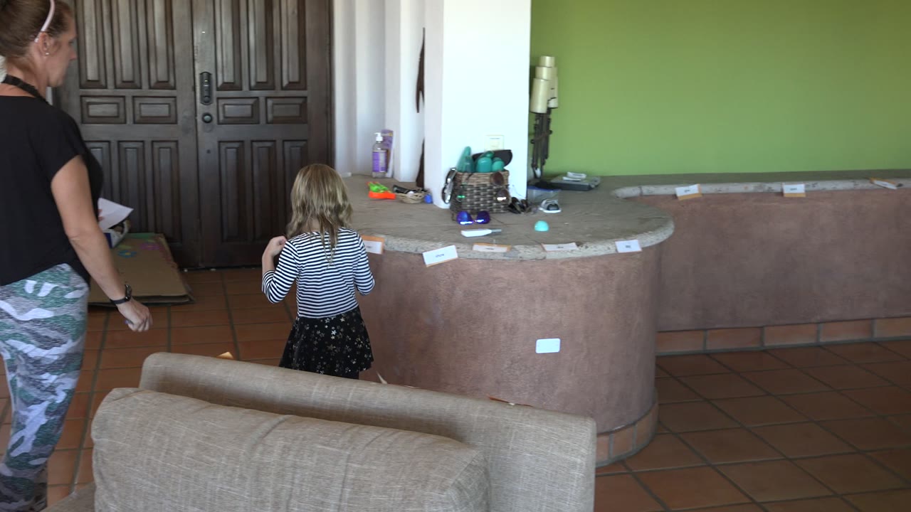 In Los Cabos, Mexico, the child faces the curved counter with small paper cards spread along the edge as the woman stands behind her in the tiled indoor space.