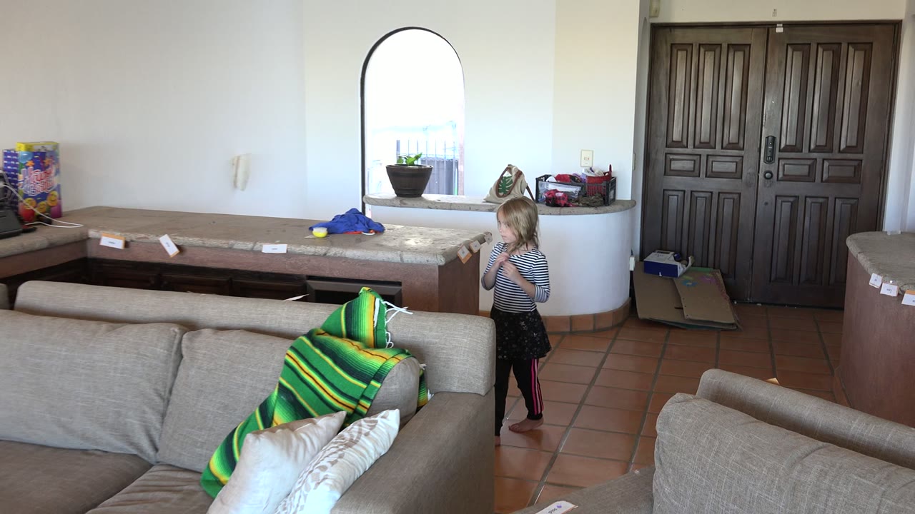 A young girl stands beside a tan couch in a Los Cabos home interior, with a long counter, arched window, pillows, and dark double doors behind her.