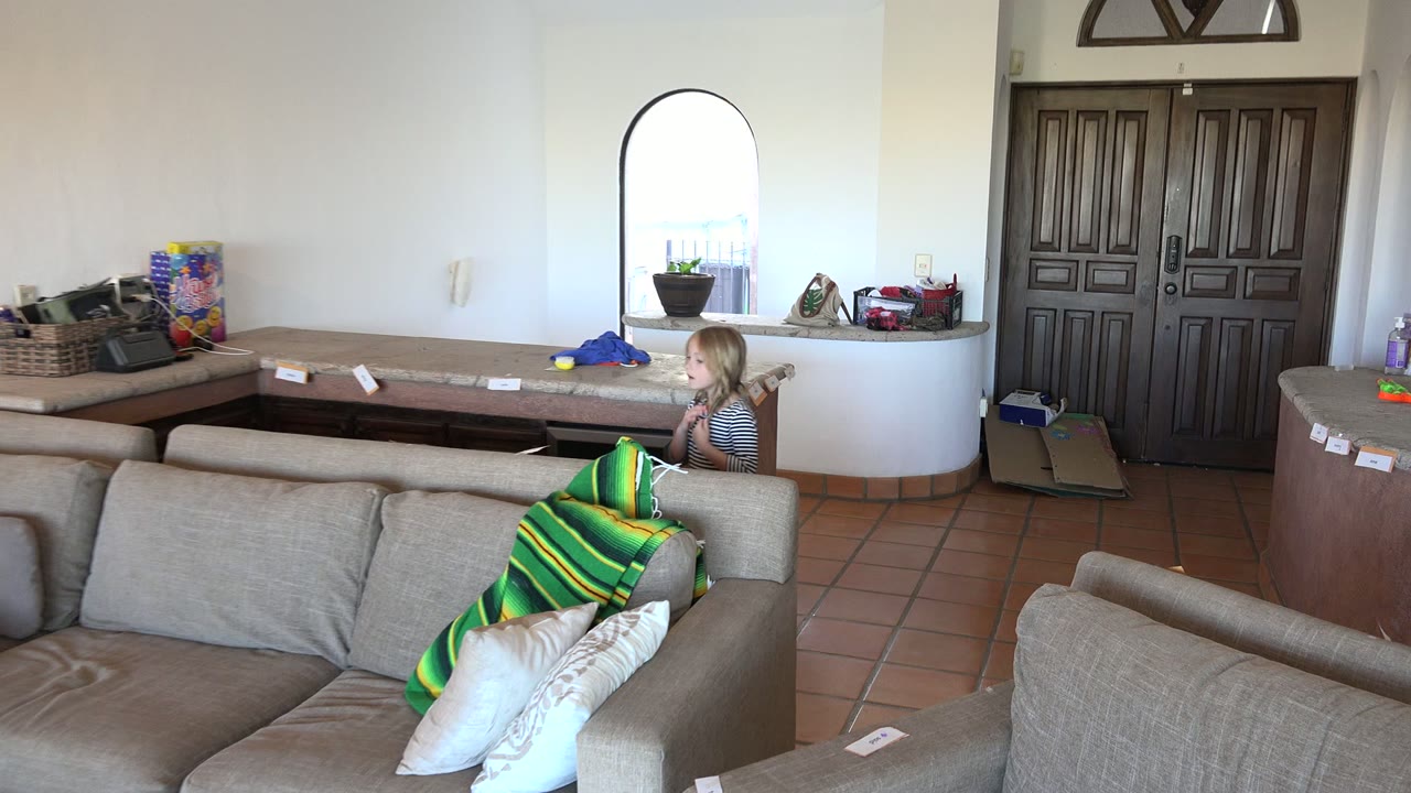 The girl has moved farther right behind the couch in the Los Cabos room, with the arched window and a cluttered counter visible across the back wall.
