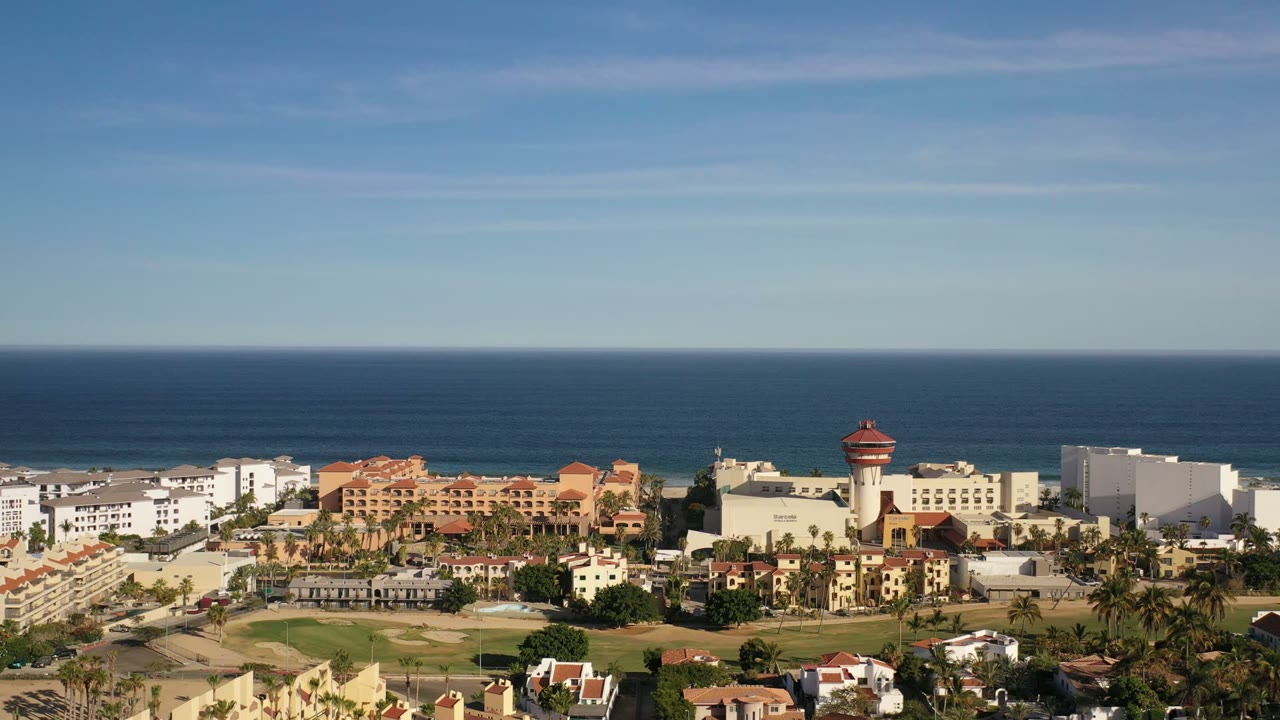 Second frame shows the Los Cabos waterfront property area from above, with a red-roofed tower feature, white hotel structures, palms, and no visible people.