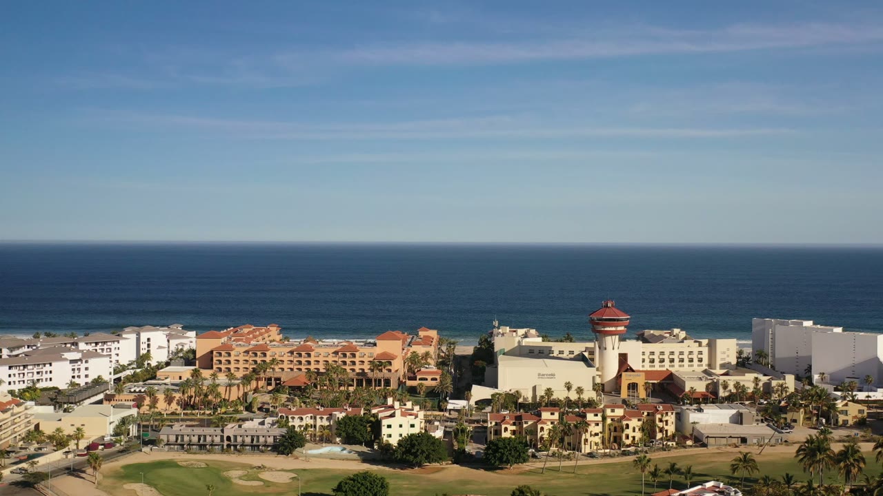 Aerial view in Los Cabos, Mexico, showing beachfront hotel blocks, palm rows, a golf course edge, sandy shoreline, dark blue ocean, and no visible people.