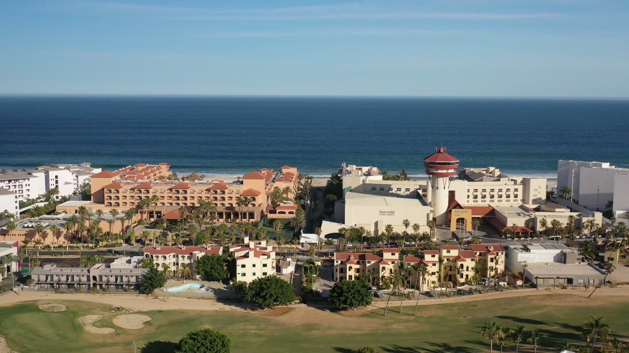 Los Cabos aerial frame with a red-roofed tower near the right side, low-rise hotel and apartment buildings across the center, palm rows along roads, golf course below, ocean behind, and no visible people.