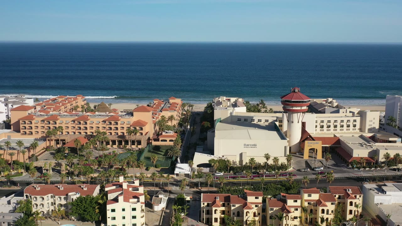 Los Cabos aerial frame with the ocean horizon, narrow white surf, cream resort buildings, orange roof sections, palm-lined road, foreground residential-looking blocks, and no visible people.