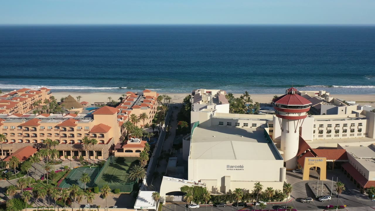 Aerial frame over a Los Cabos beachfront resort with a red-roofed tower, pale hotel blocks, palm landscaping, sport courts, road, blue ocean, and no visible people.