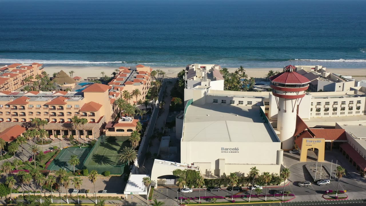 Drone view in Los Cabos showing the resort tower nearer the center, terracotta roofs to the left, white buildings, palms, beach, ocean bands, and no visible people.