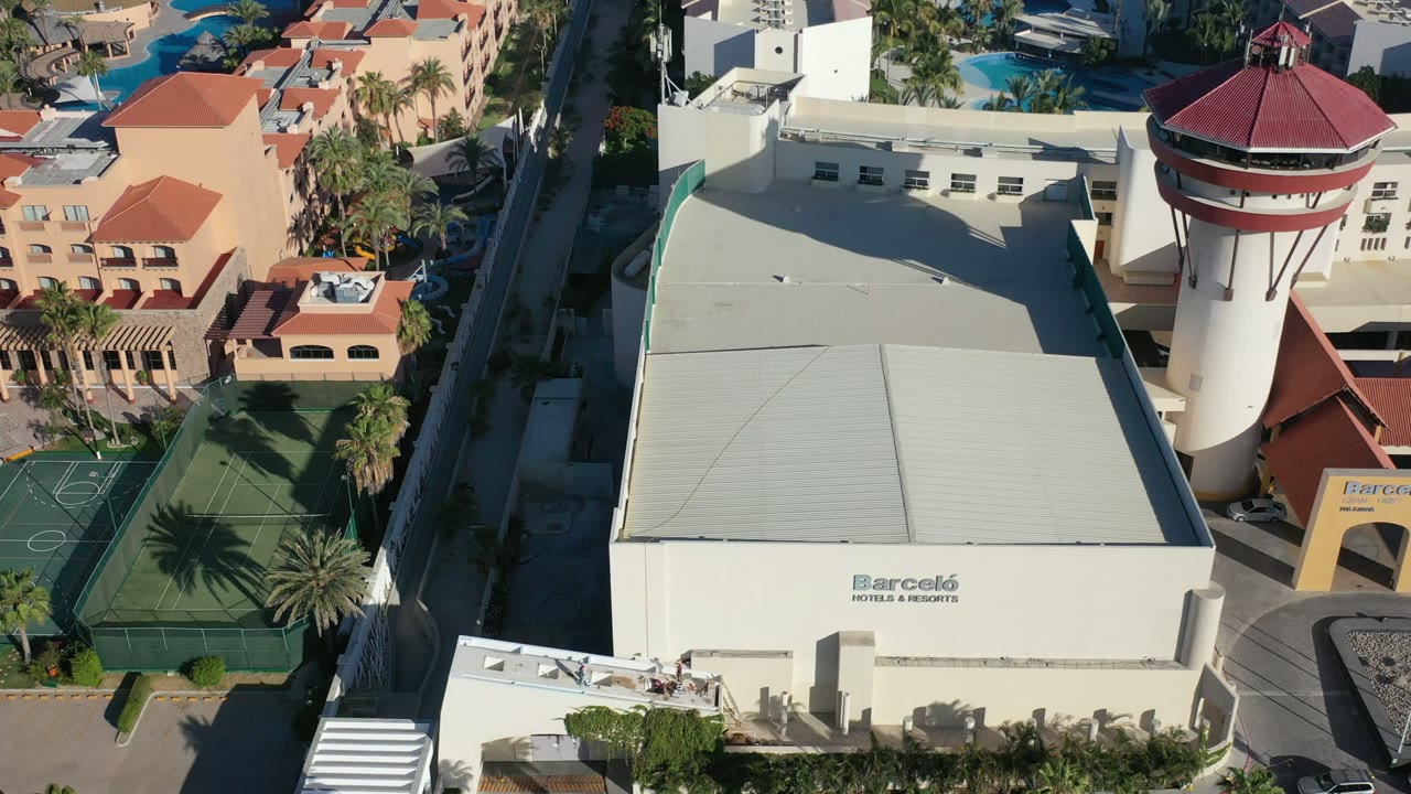 Aerial frame over a Los Cabos resort-style building with a broad pale roof, white tower with red cap, tennis courts, palms, blue pools, and no visible people.