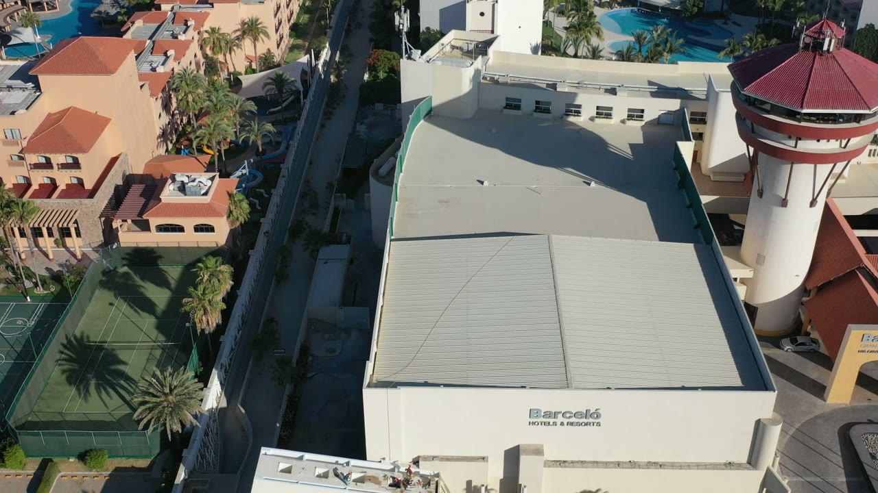 High aerial view in Los Cabos with the resort roof centered, the red-roofed tower to the right, tennis courts below, pools behind, and no visible people.