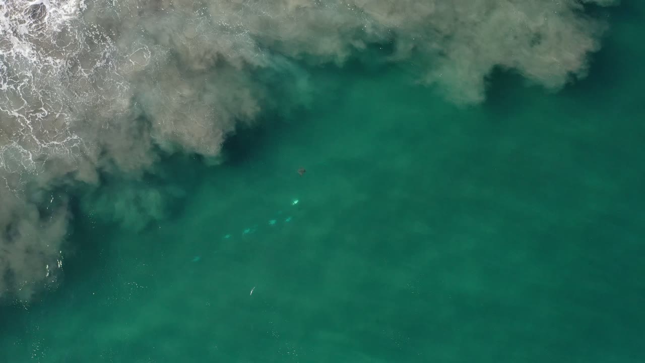 Aerial view over Los Cabos ocean water, with surf foam along the left edge, gray cloudy water near the top, small bright turquoise patches, and no visible people.