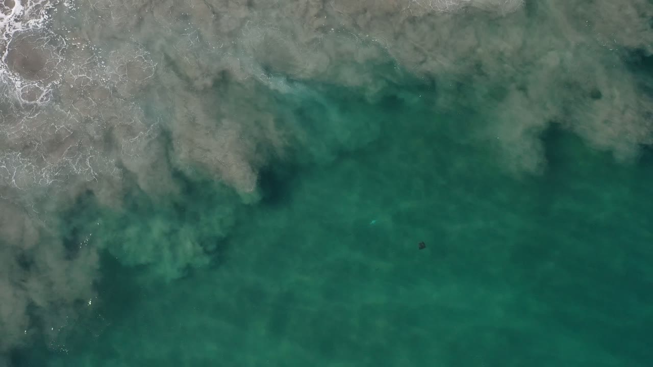 Turquoise-green water in Los Cabos, Mexico, seen from above as cloudy wave foam spreads across the upper frame with no visible people.