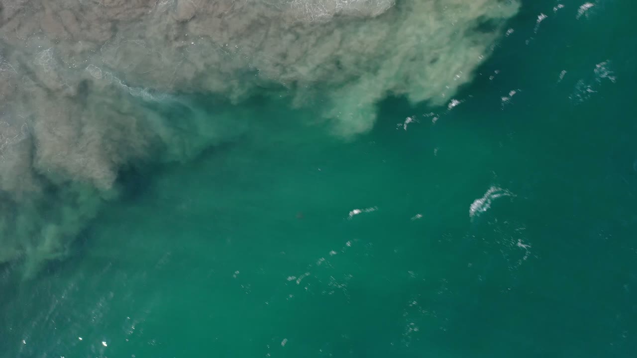 Aerial view over green-blue water in Los Cabos, Mexico, with a pale shoreline edge at the top left, scattered white wave breaks, and no visible people.