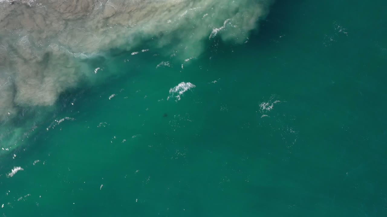 Aerial Los Cabos water frame with the pale coastal edge across the upper left, green sea filling the image, small whitecaps, and no visible people.