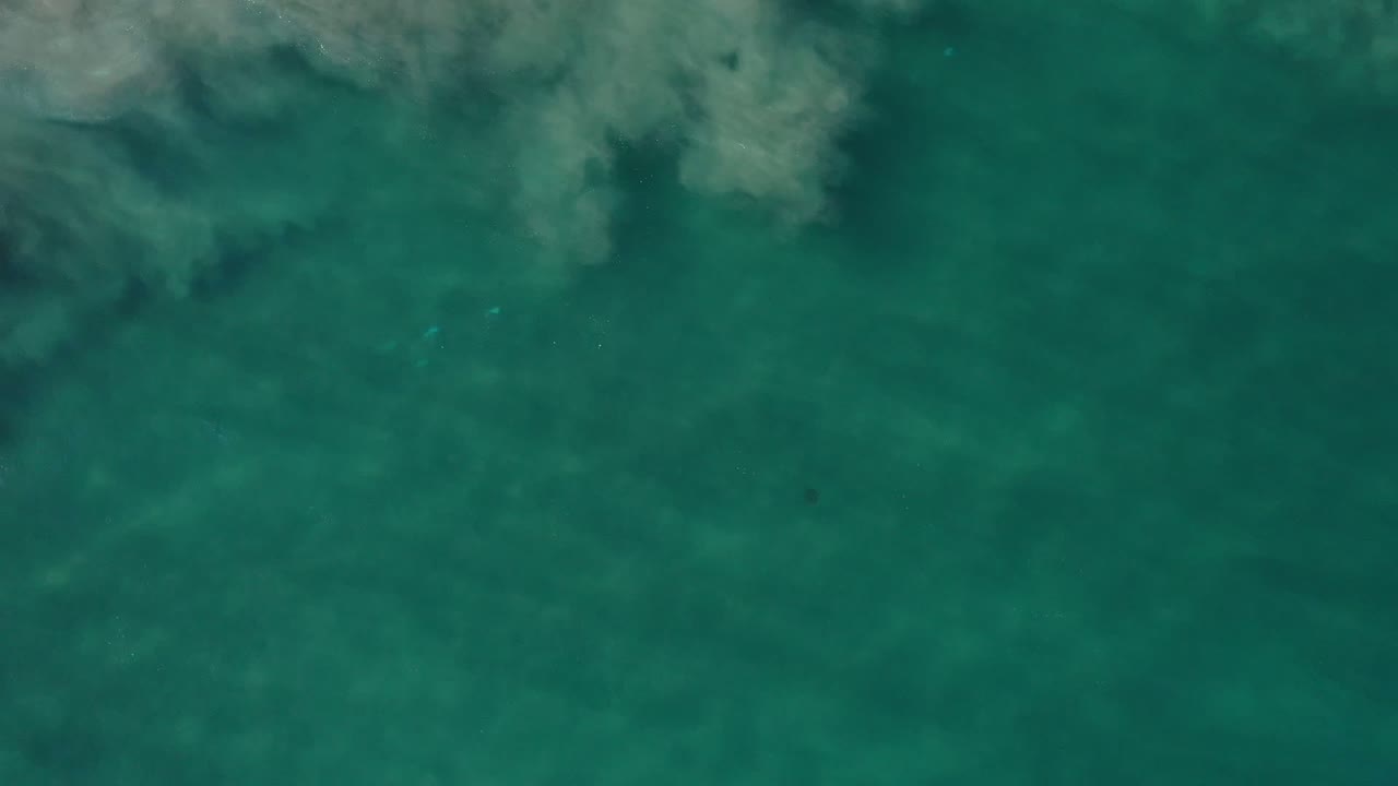 Blue-green shallow seawater in Los Cabos, Mexico, with cloudy pale sand or foam patterns spread across the upper part of the frame and no visible people.