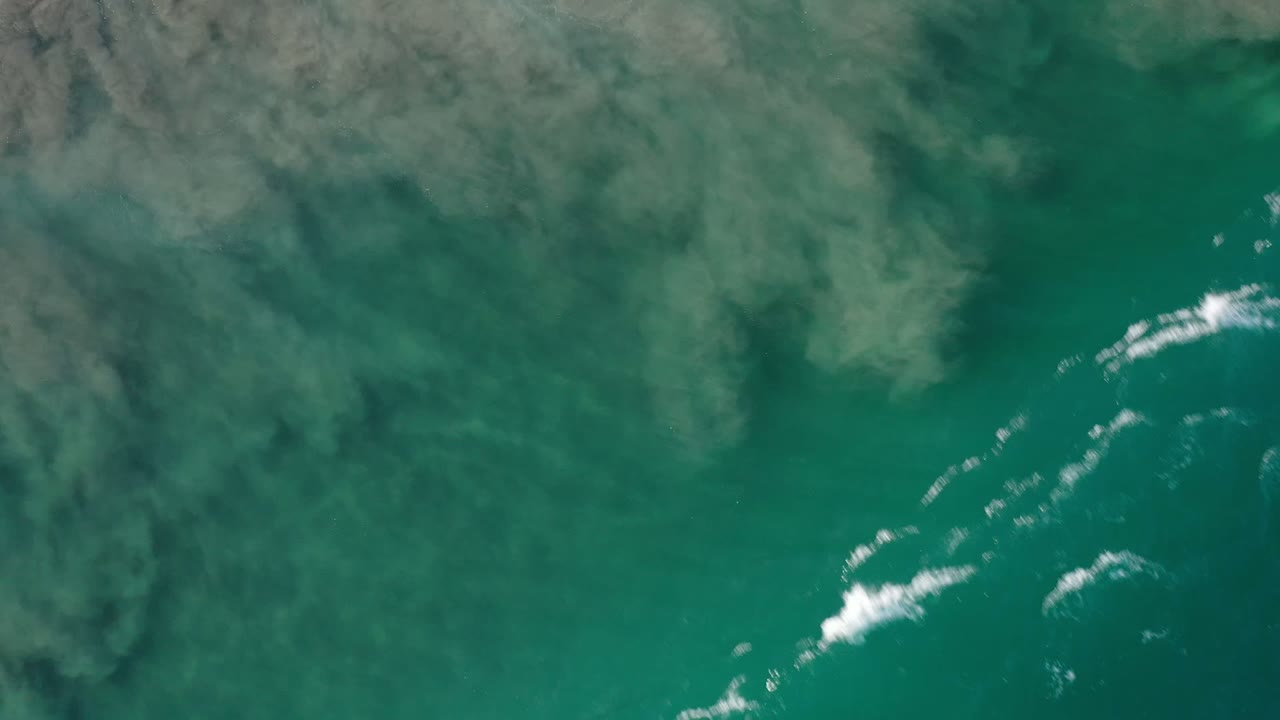 Aerial Los Cabos ocean view with teal water, muted shoreline texture, and a narrow white surf break starting at the right edge, with no visible people.