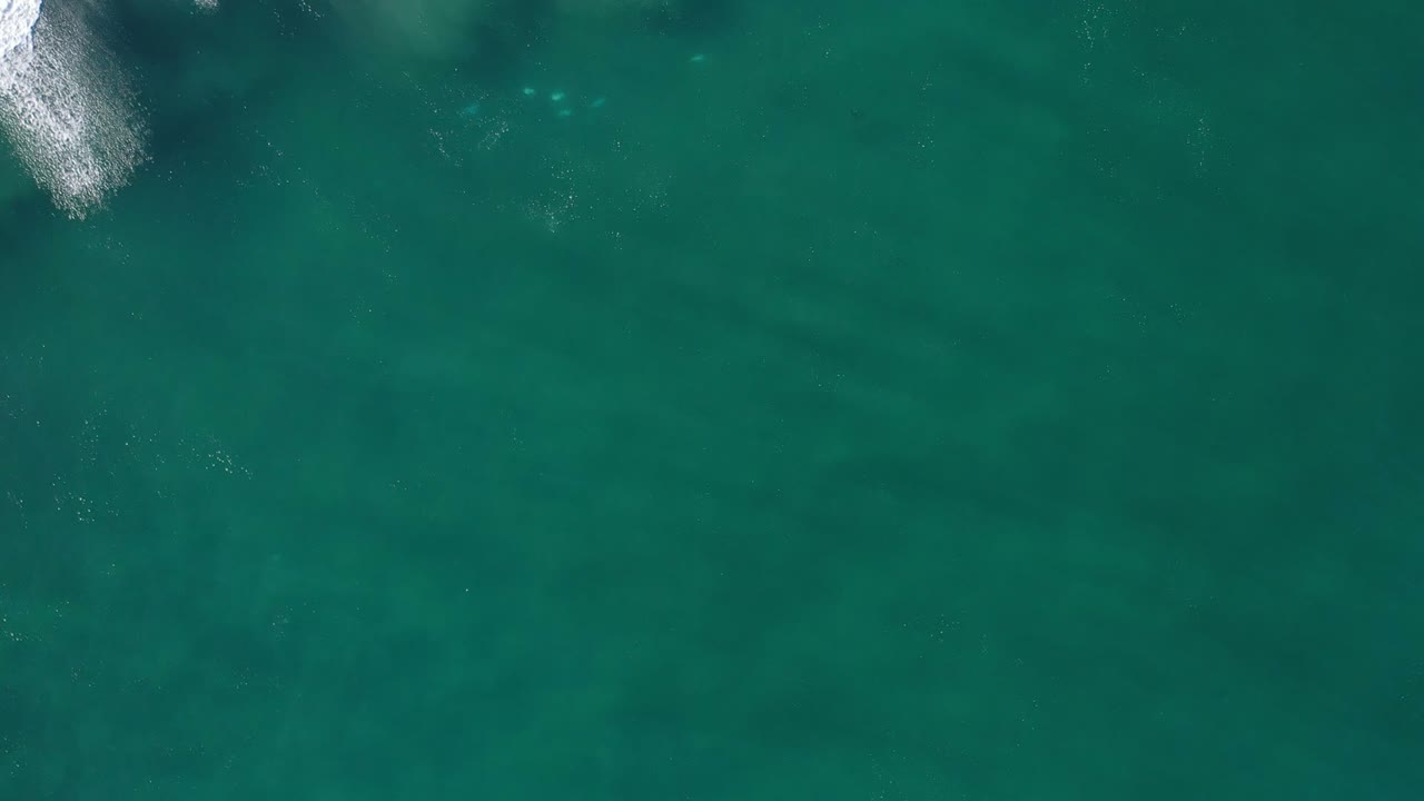 Aerial view of clear blue-green water in Los Cabos, Mexico, with a small white breaking wave near the upper edge and no visible people.