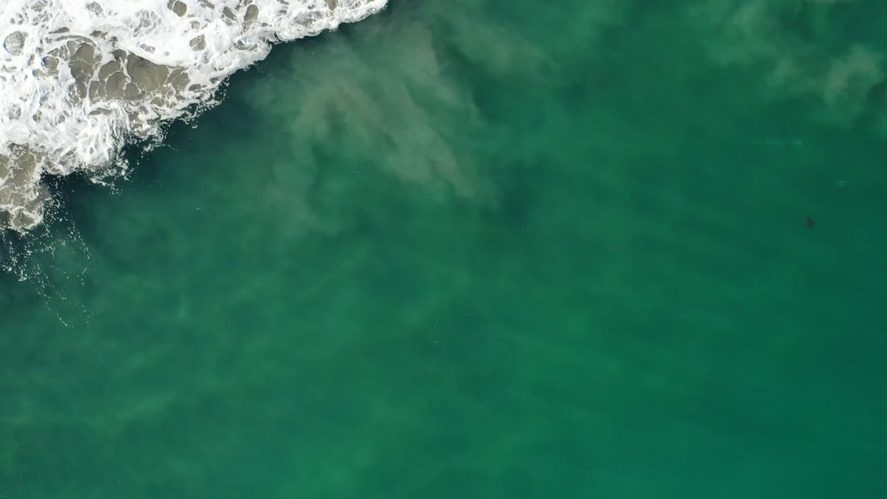 Aerial top-down frame from Los Cabos, Mexico showing a thicker band of white wave foam pushing over green water, with no visible people.
