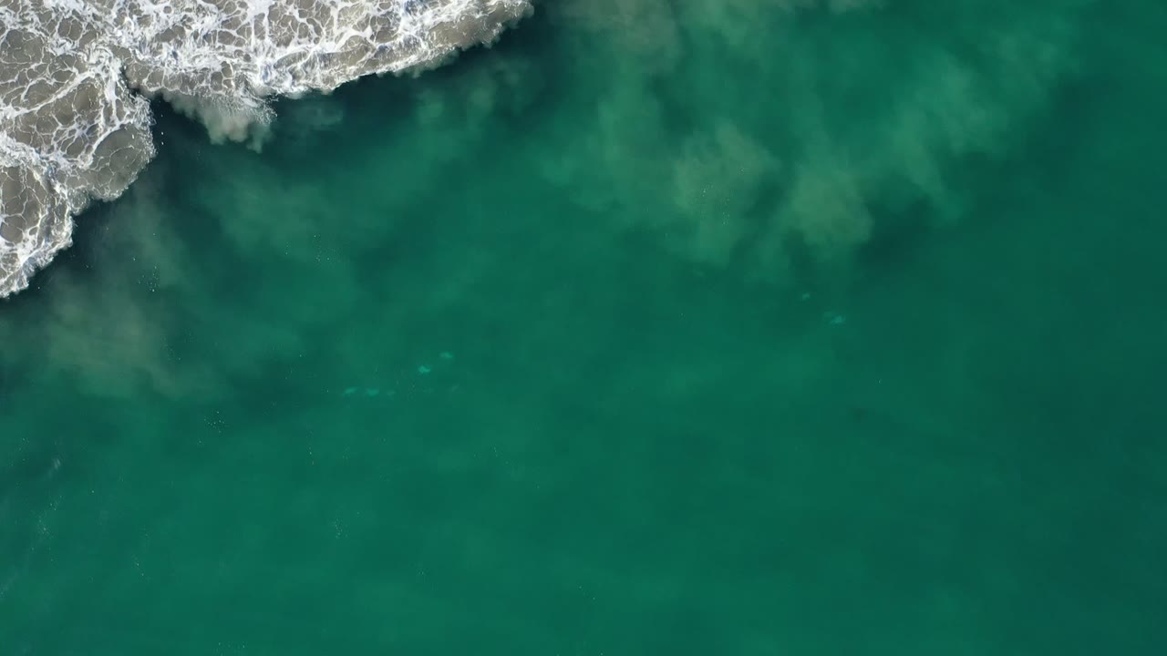 Overhead teal ocean with white surf breaking on rocky shoreline.