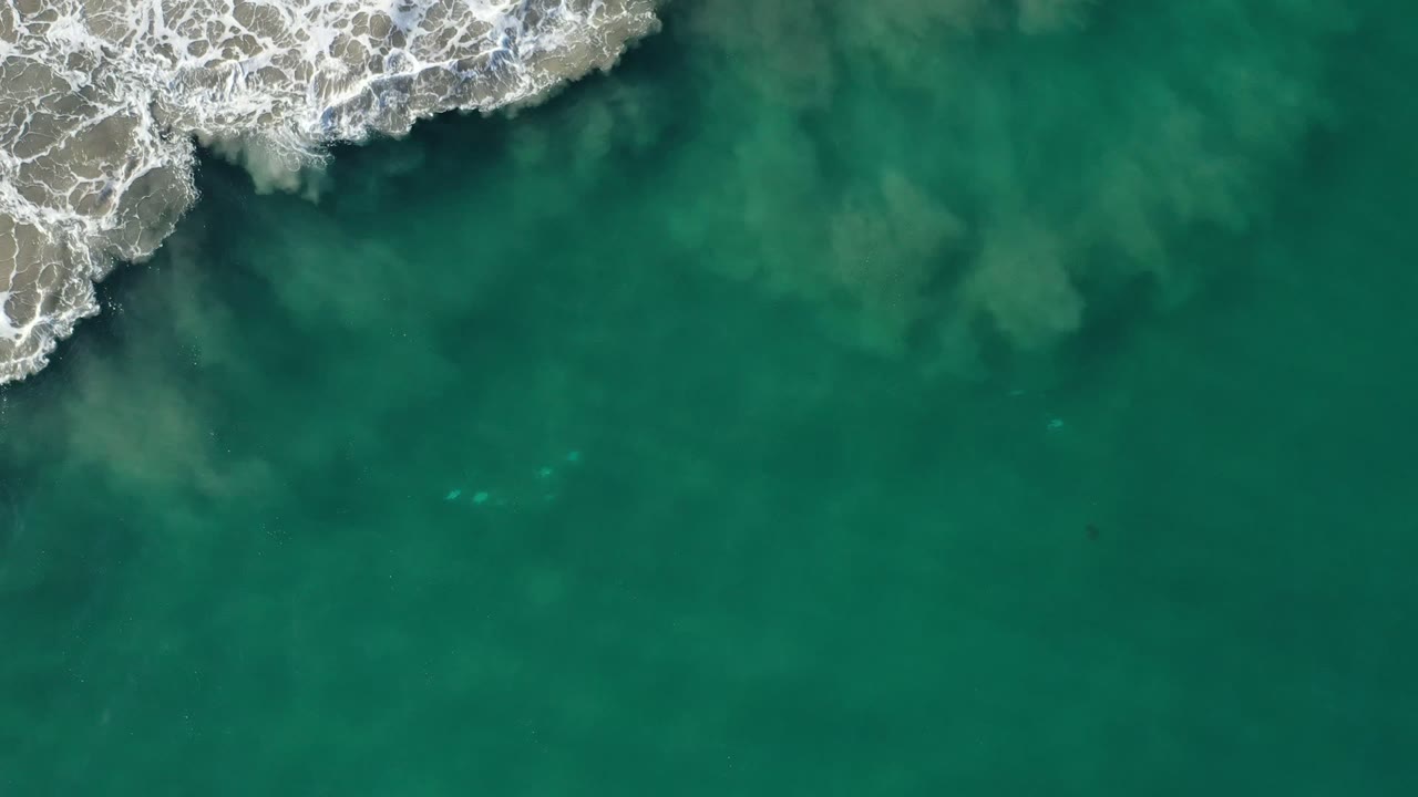 Overhead Los Cabos frame showing a rock point at the upper left, breaking white water around it, and broad green-blue ocean below, no visible people.