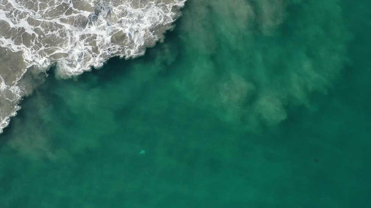 Overhead Los Cabos, Mexico beach frame with white surf entering from the upper left and green water filling the rest of the view, with no visible people.