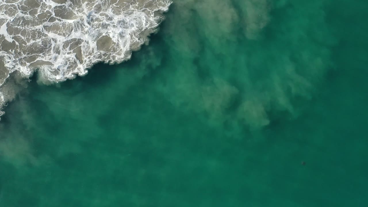 Overhead Los Cabos, Mexico view with foamy surf near the top edge and cloudy sand movement visible below the green ocean surface, with no visible people.